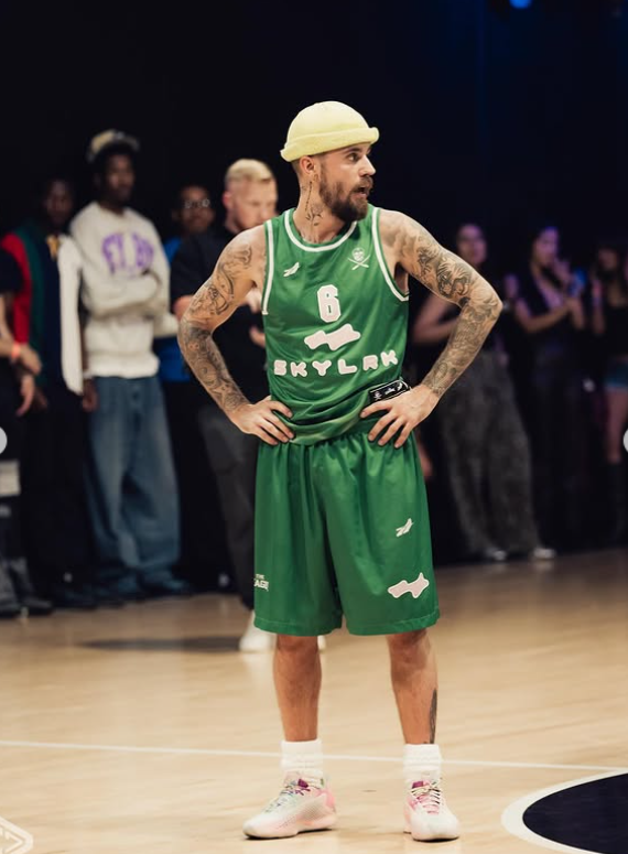 Basketbalista v zelenom drese a prilbe stojí na palubovke, obklopený divákmi v pozadí.