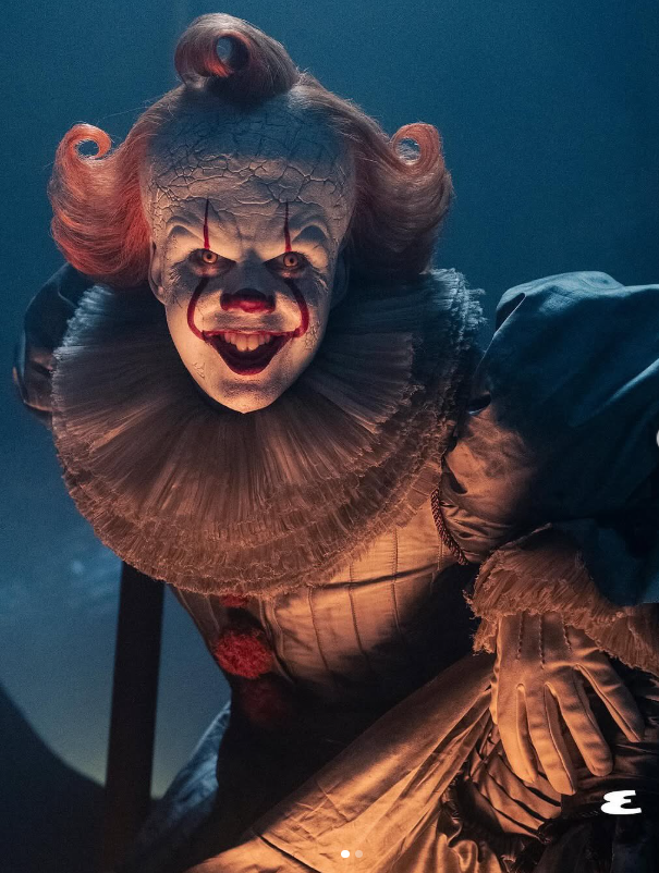 Strašidelný klaun s červeným nosom a rúžovými vlasmi usmievajúci sa do kamery v tmavej atmosfére.