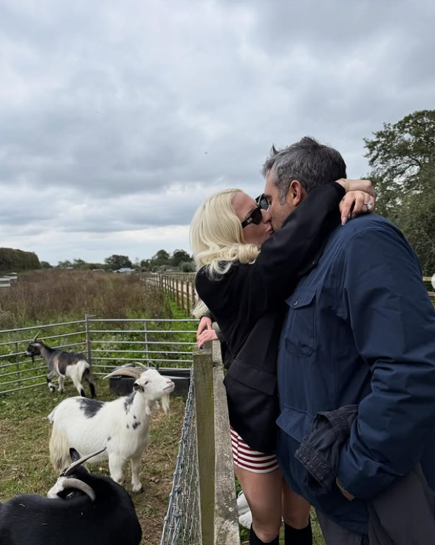 Pár sa bozkáva pri ohrade s kozami na farme pod zamračenou oblohou.