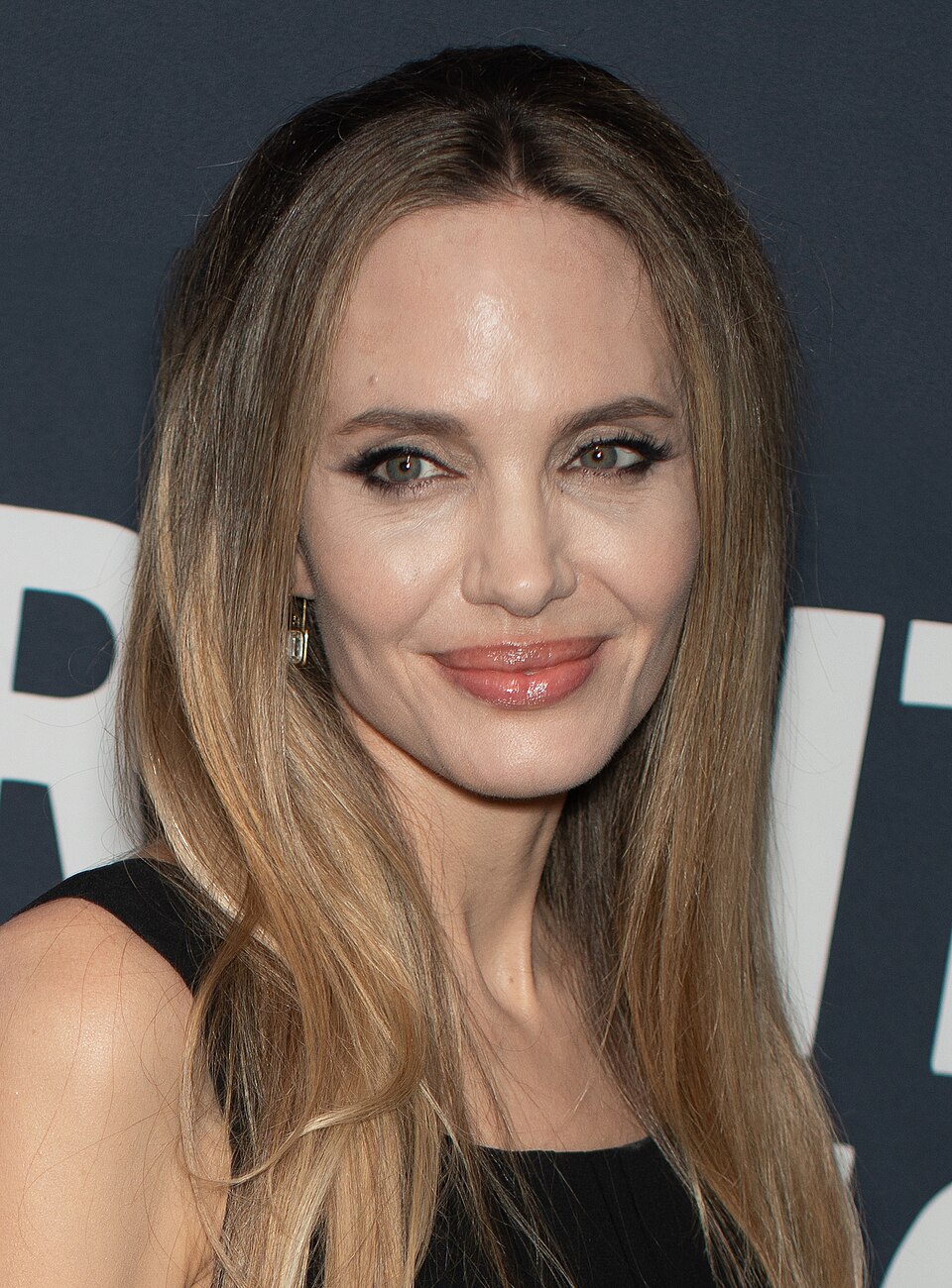 Osoba s dlhými vlasmi v čiernom oblečení, úsmev na tvári, pózuje na červenom koberci pri filmovej premiére.