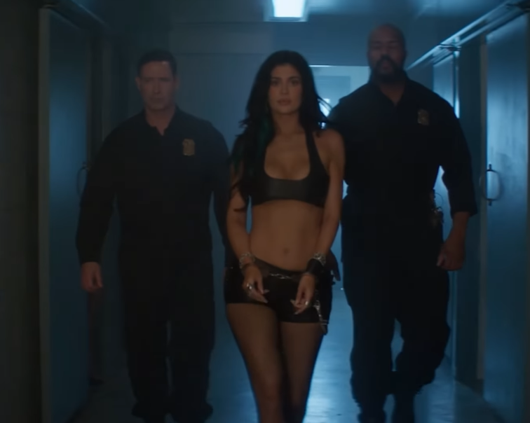 Osoba v čiernom outfite kráča chodbou po boku dvoch mužov v uniformách.