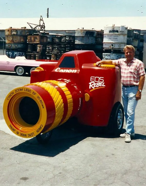 Veľký model fotoaparátu Canon EOS Rebel na kolieskach so stojacim mužom vonku.