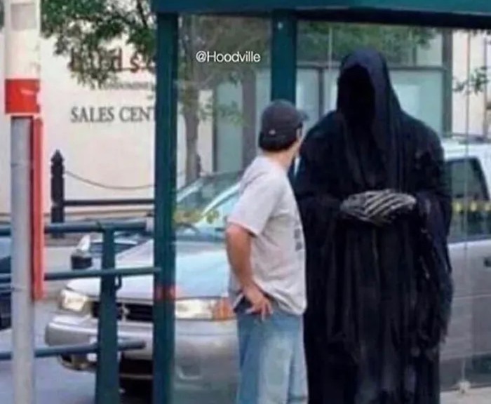 Osoba stojí na zastávke a vedľa nej stojí postava v čiernom plášti, pripomínajúca smrť.