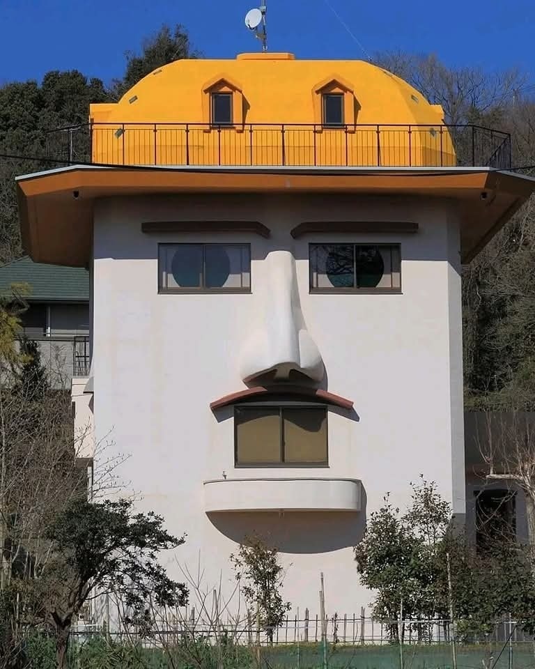 Dom so žltou strechou a fasádou, ktorá pripomína tvár s očami, nosom a ústami v lese.