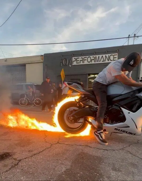 Motocyklista na Suzukim predvádza ohnivý burnout pred dielňou.