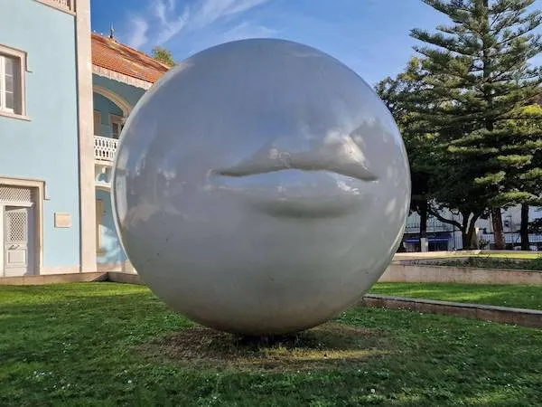 Veľká biela guľa so sochou pier na trávniku pred modrou budovou a stromami.
