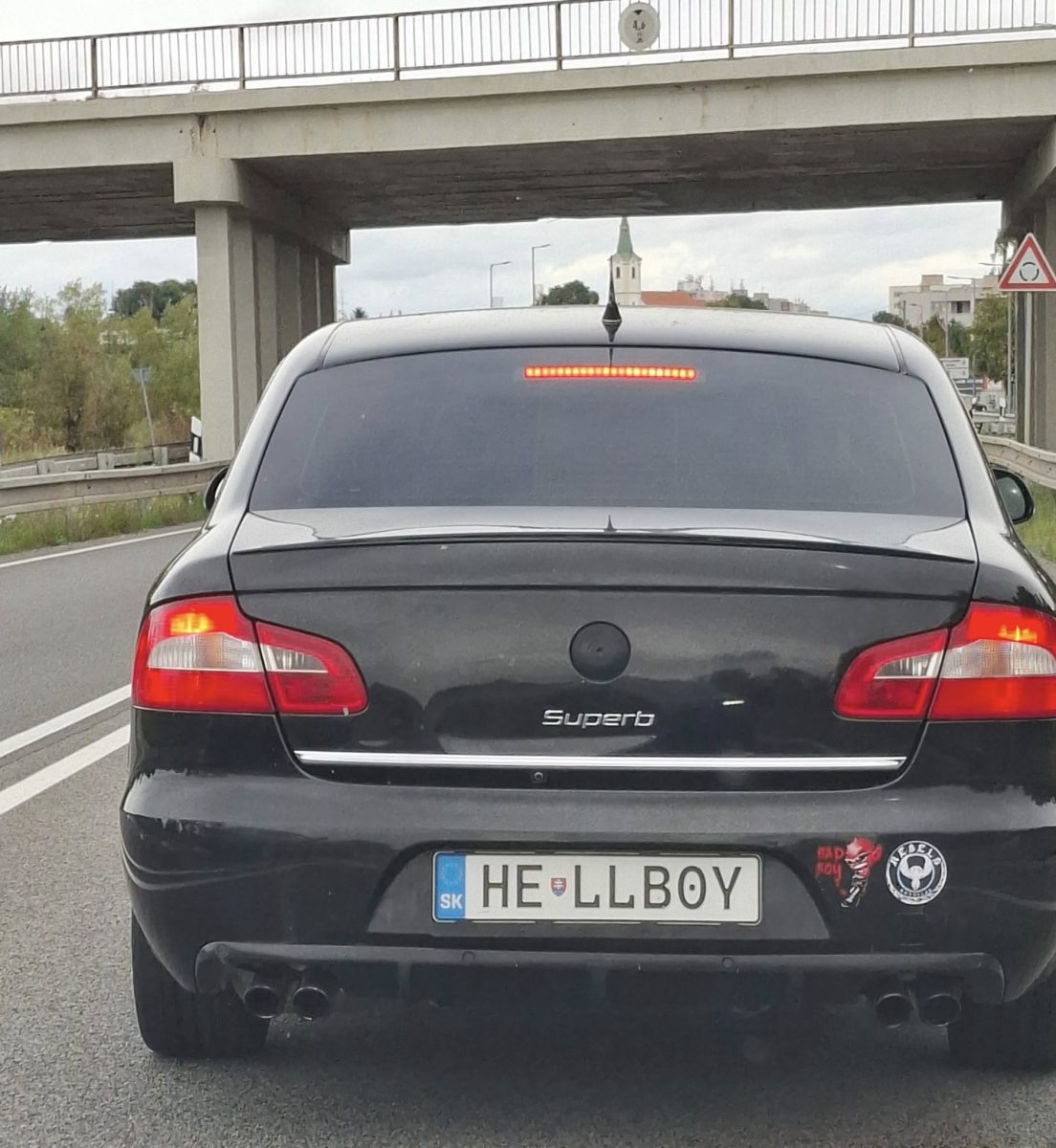 Čierne auto so špeciálnou poznávacou značkou HEL-LBOY na ceste pod mostom, s nálepkami na nárazníku.