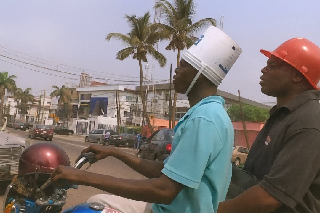 Dve osoby na motorke s improvizovanými helmami, palma a mestská ulica v pozadí.