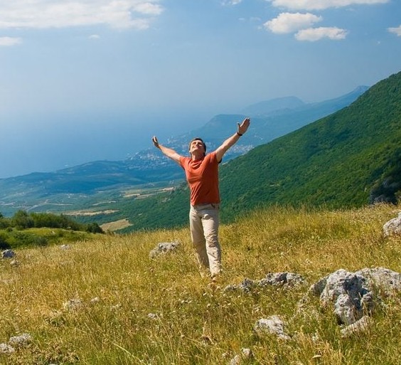 Osoba s roztiahnutými rukami stojí na horskej lúke s výhľadom na more a okolitú prírodu, vyjadrujúc pocit slobody.
