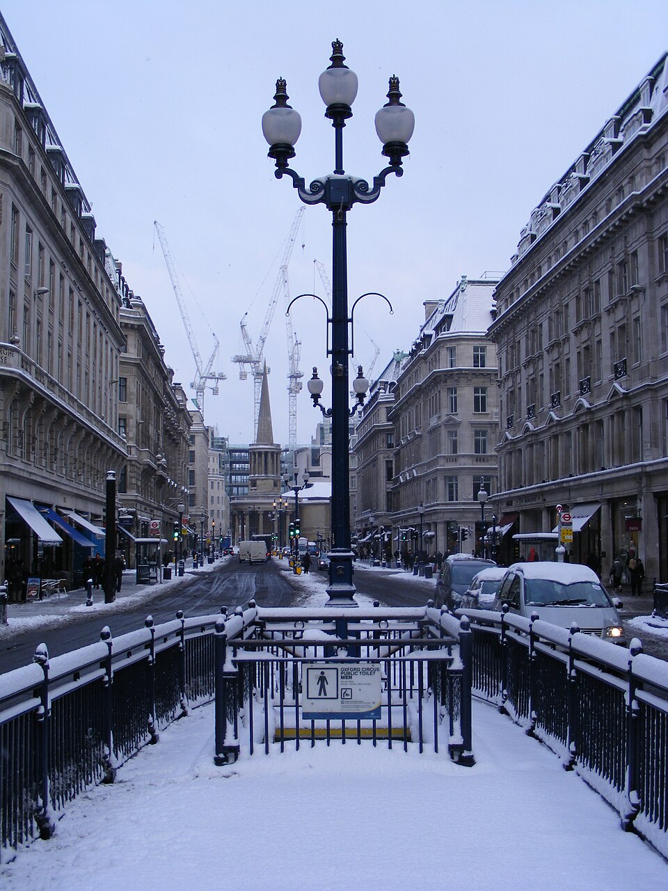 Zasnežená ulica v meste s historickými budovami a lampou uprostred, v pozadí žeriavy a zima.