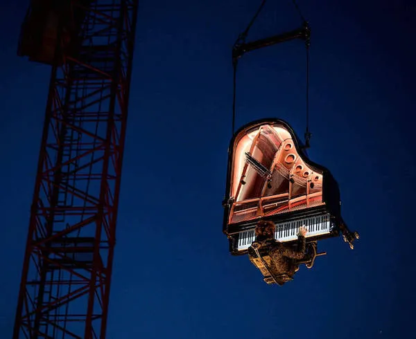 Klavír zavesený na žeriave v noci, osvetlený zvnútra, vytvára dramatickú scenériu na tmavej oblohe.