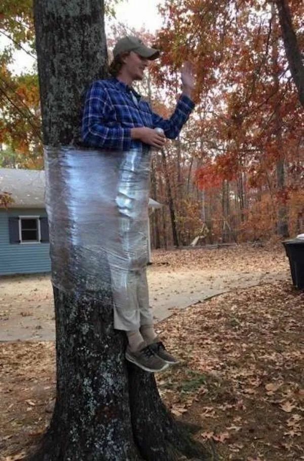 Osoba oblečená v károvanej košeli pripevnená na strom fóliou, jesenné lístie v pozadí.