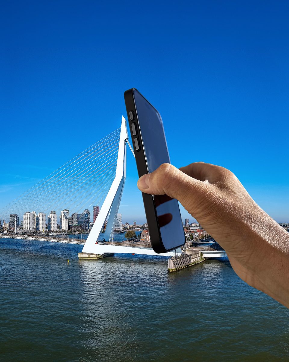 Ruka držiaca telefón pri fotografovaní moderného mosta nad vodnou plochou pod modrou oblohou.