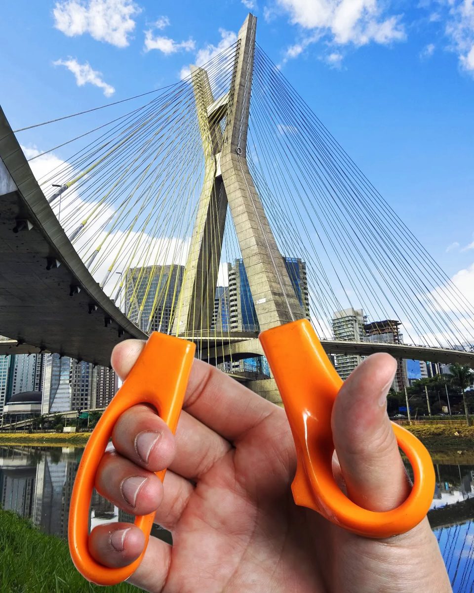 Ruka drží oranžové nožnice pred moderným lanovým mostom pod modrou oblohou.