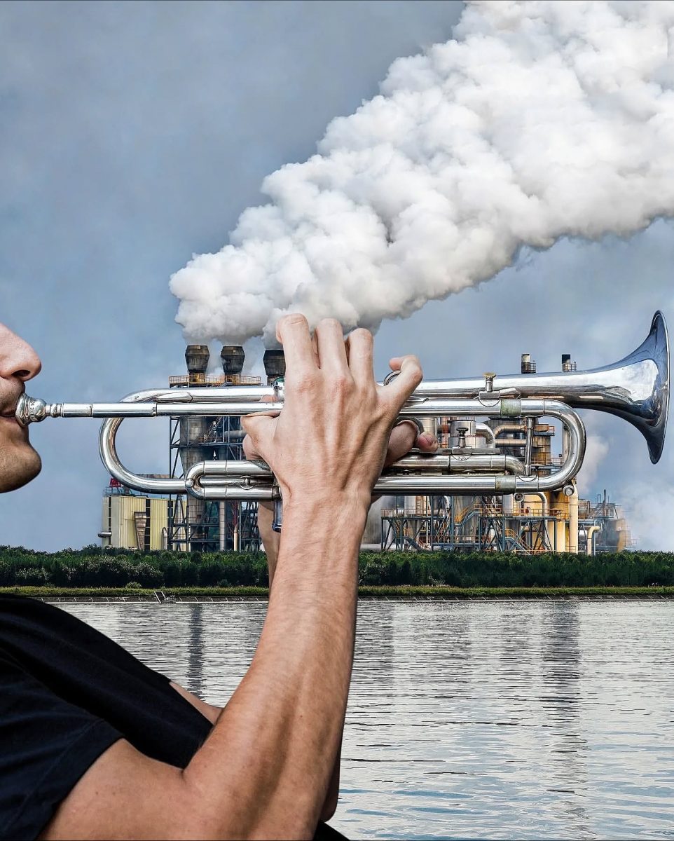 Osoba hrá na trúbke pri vodnej ploche, pozadie tvorí továreň vypúšťajúca dym, tvorba dymu vrcholí z trúbky do oblakov.