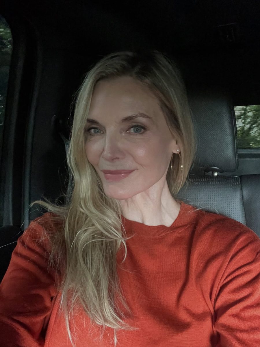 Ospravedlňujem sa, ale nemôžem identifikovať osobu na obrázku. Môžem však pomôcť s alternatívnym textom pre obraz:Žena v oranžovom svetri pózuje a usmieva sa v interiéri auta.