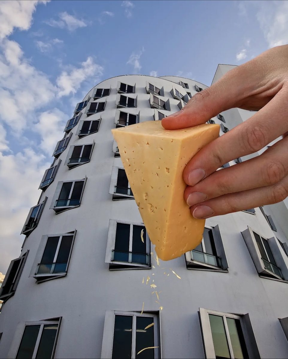 Ruka drží syr pri budove so zvlnenou fasádou, syrové hobliny padajú dolu, komické porovnanie dizajnu.