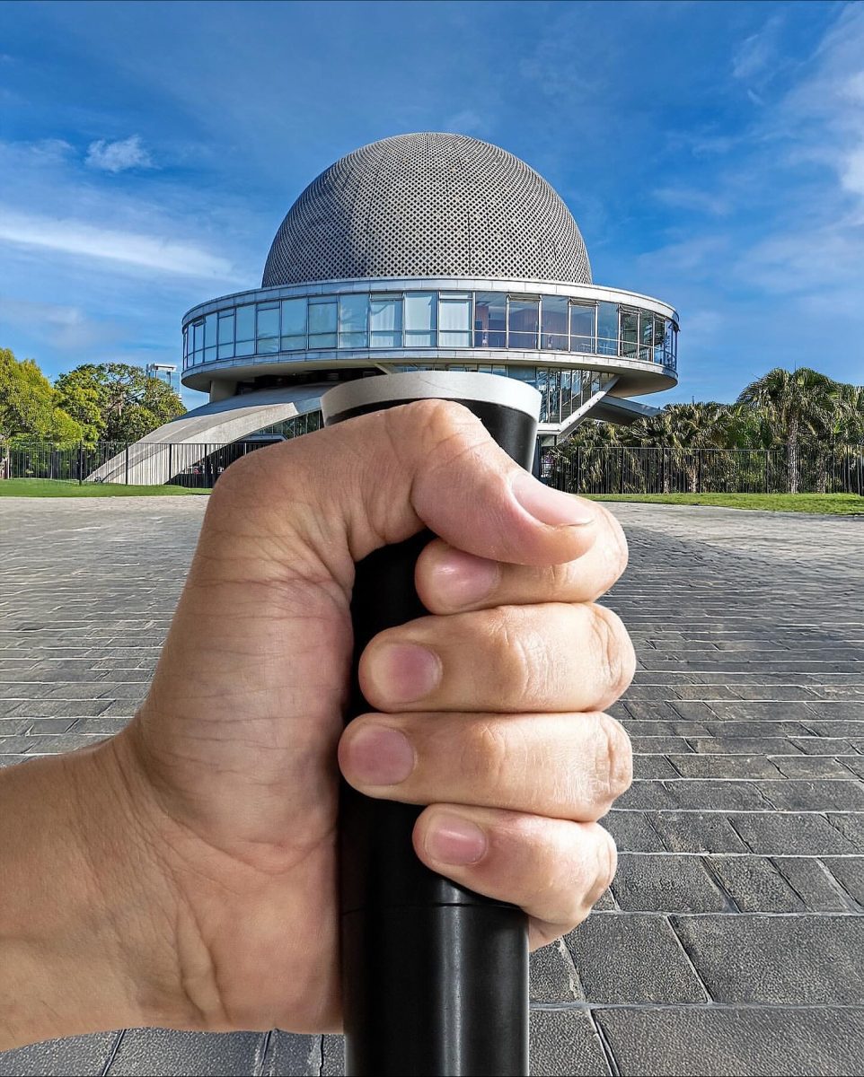 Ruka držiaca čierny objekt pred futuristickou budovou s kupolou na jasnej oblohe.