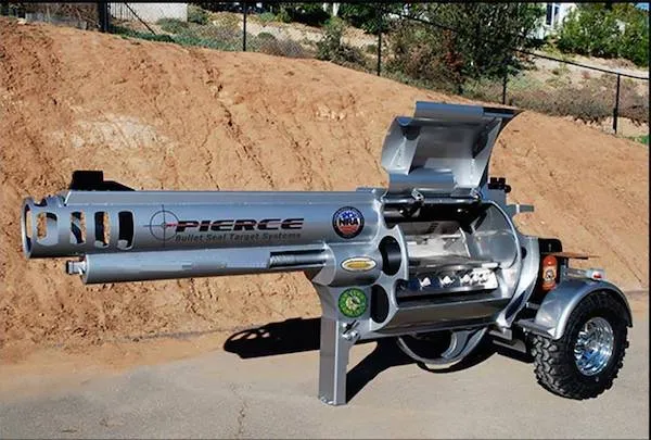 Veľký gril v tvare revolvera na kolieskach postavený na spevnenom podklade pred hlineným kopcom.