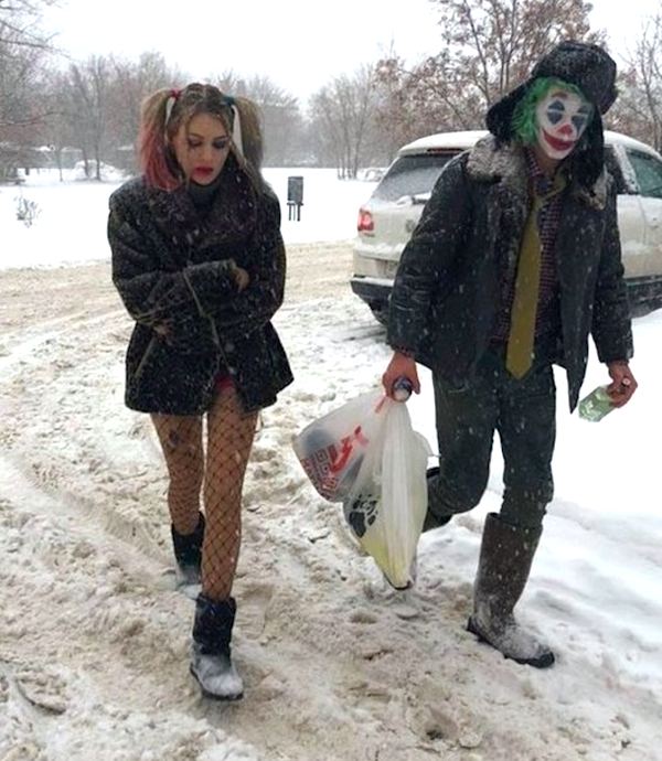 Dvaja ľudia v kostýmoch klauna a Harlekýna kráčajú zasneženou cestou s nákupnými taškami v ruke.