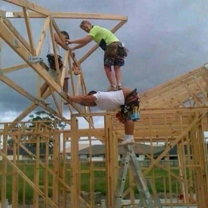 Stavbári balansujúci na improvizovanom lešení, konštrukcia dreveného domu so zamračenou oblohou v pozadí.