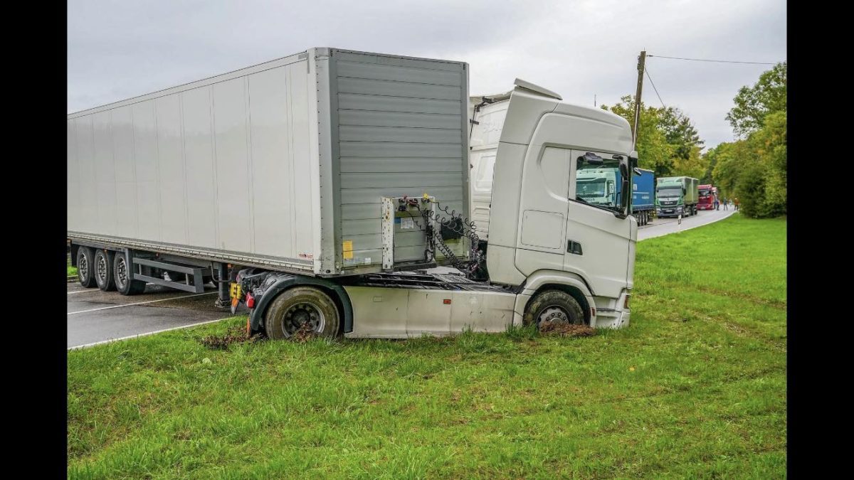 Kamión zaseknutý na tráve vedľa cesty, s ostatnými vozidlami v pozadí.