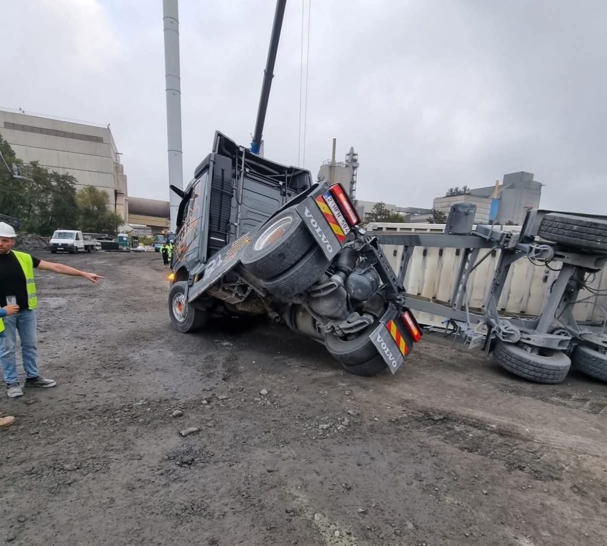 Prevrátený kamión na stavenisku s vykladačom v pozadí a pracovníkmi v reflexných vestách.