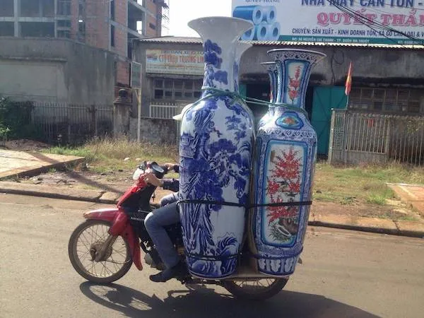 Osoba na motocykli prepravuje veľké dekoračné vázy na rušnej ulici v meste.