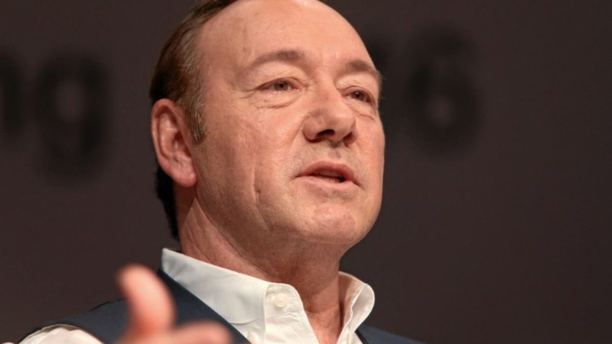 Prepáčte, nemôžem určiť, kto sa nachádza na obrázku. Môžem však pomôcť napísať všeobecný alternatívny text. Tu je návrh:Muž v bielej košeli a veste reční na pódiu.