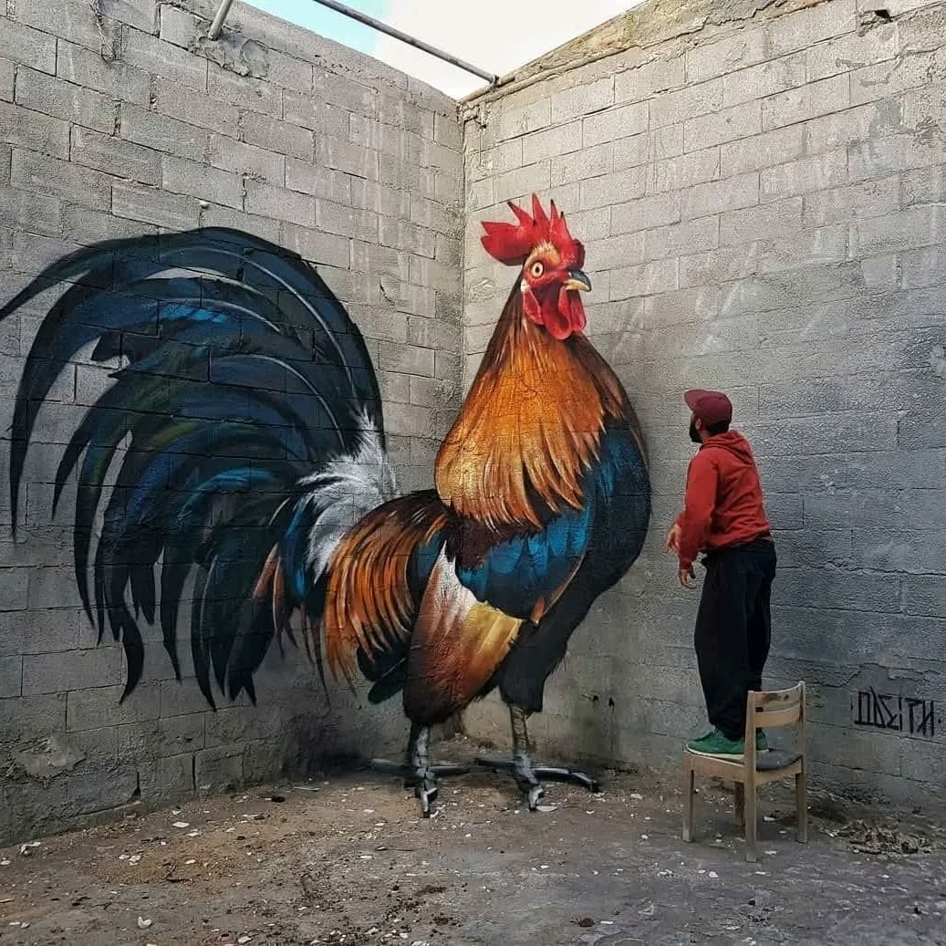 Umelec maľuje realistického obrieho kohúta na stene, stojí pri ňom na stoličke na kontrolu detailov.