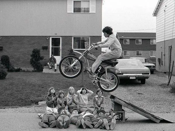 Chlapec na biku skáče cez rampu nad skupinou detí ležiacich na ceste pred domom.