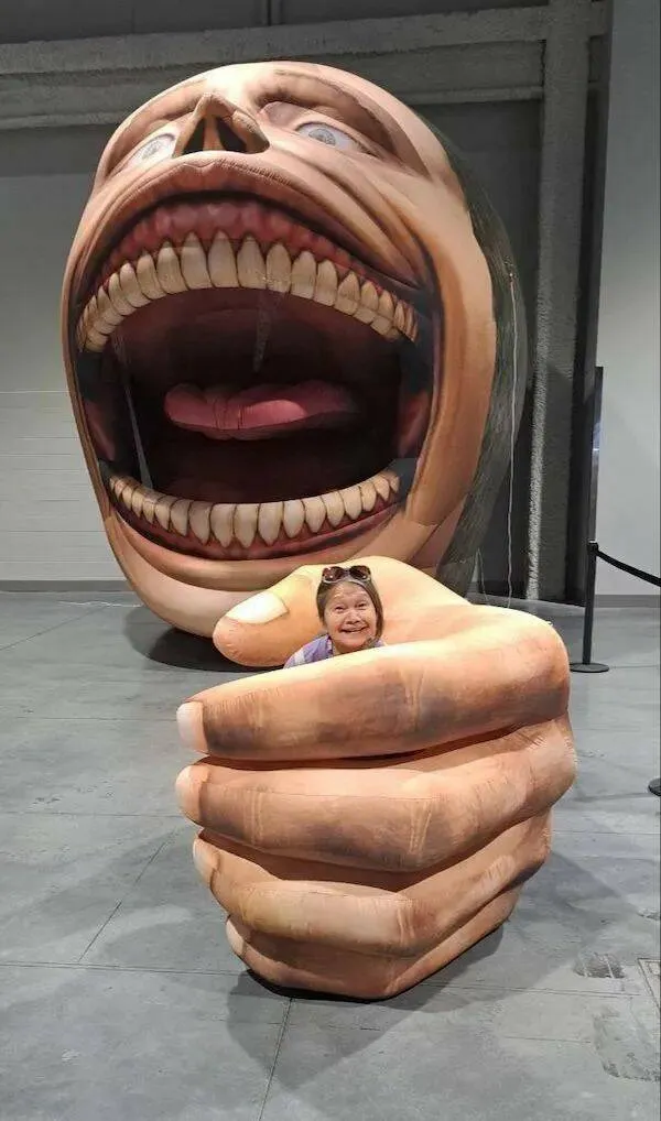 Staršia žena sedí v obrovskej sochárskej ruke pred hlavou so široko otvorenými ústami v galérii.