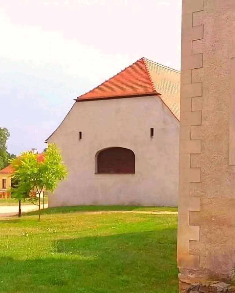 Biela budova s červenou strechou na trávnatom pozemku, vedľa stromu, slnečný deň.