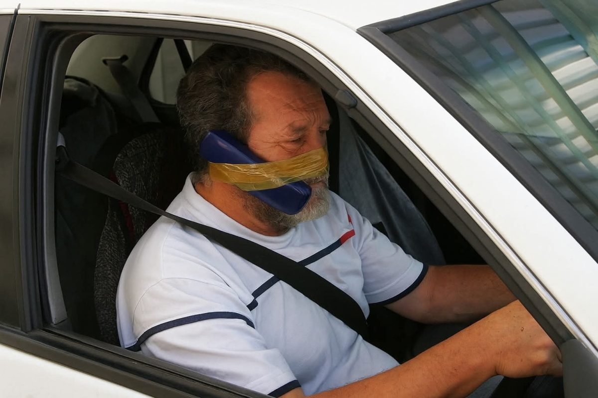 Vodič s telefónom prilepeným páskou na tvár počas jazdy autom, inovatívne využitie handsfree.