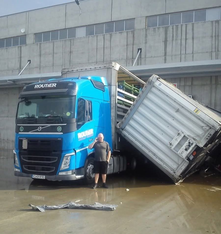 Kamión s modrým ťahačom s prevráteným návesom pri sklade, muž telefonuje.