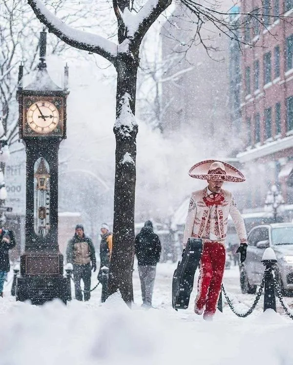 Muž v tradičnom oblečení kráča zasneženou mestskou ulicou pri hodinách.