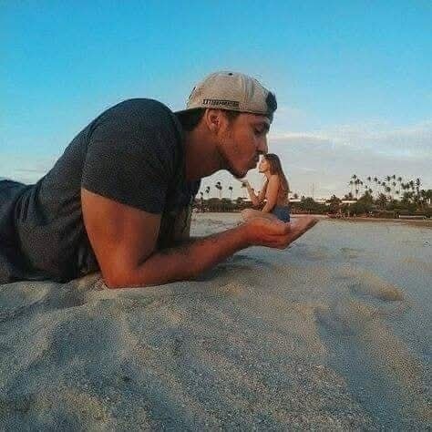 Muž na pláži drží v rukách ženu, ktorá pôsobí ako miniatúrna, vytvárajú optickú ilúziu pod žiarivým modrým nebom.