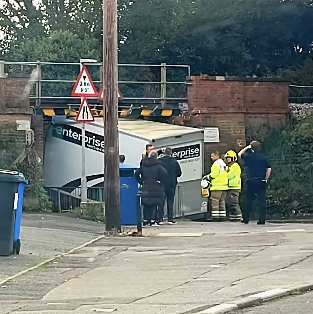 Nákladné auto zaseknuté pod nízkym mostom, okolo stoja hasiči a okoloidúci.
