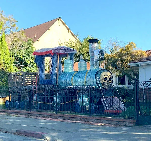 Vlaková socha z recyklovaných materiálov s lebkou na prednej časti, umiestnená v záhrade za plotom.