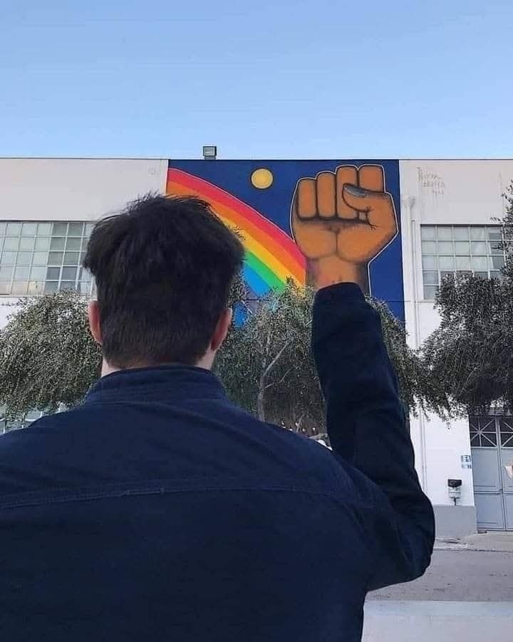 Osoba s rukou zdvihnutou pred farebnou nástennou maľbou s päsťou a dúhou na budove.