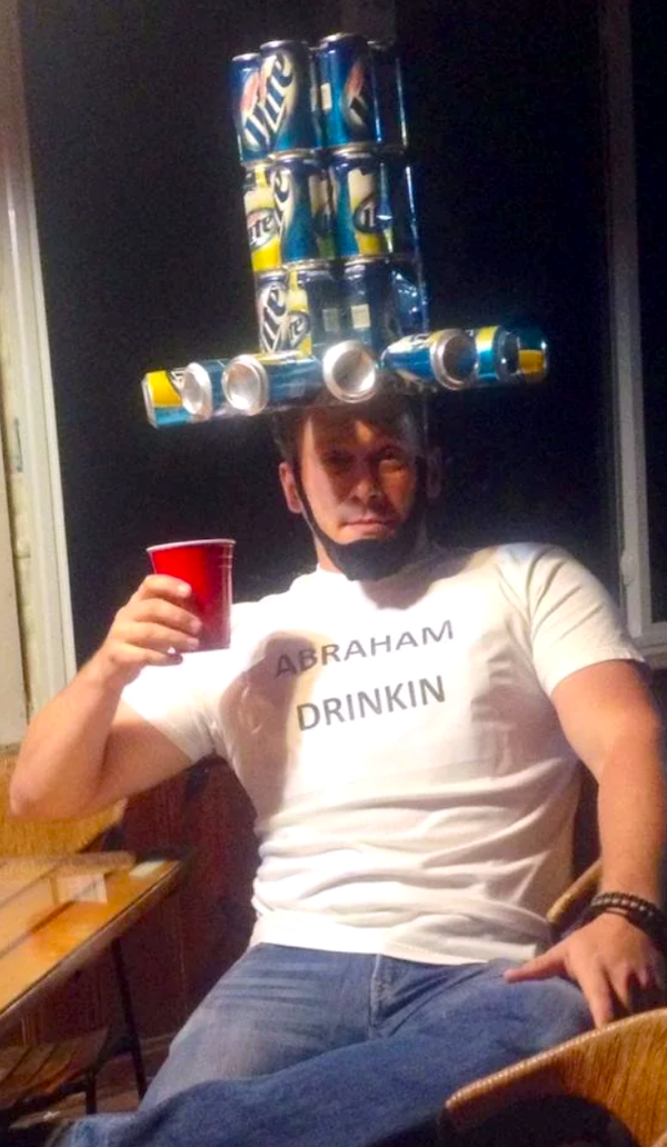 Muž s klobúkom z plechoviek a tričkom Abraham Drinkin drží červený pohár, sedí pri stole.