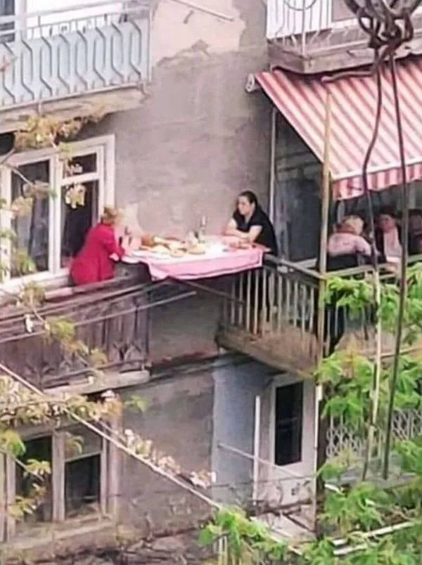 Ľudia sediaci pri stole na balkóne, zdieľajúci jedlo v mestskom prostredí, neformálne stretnutie.