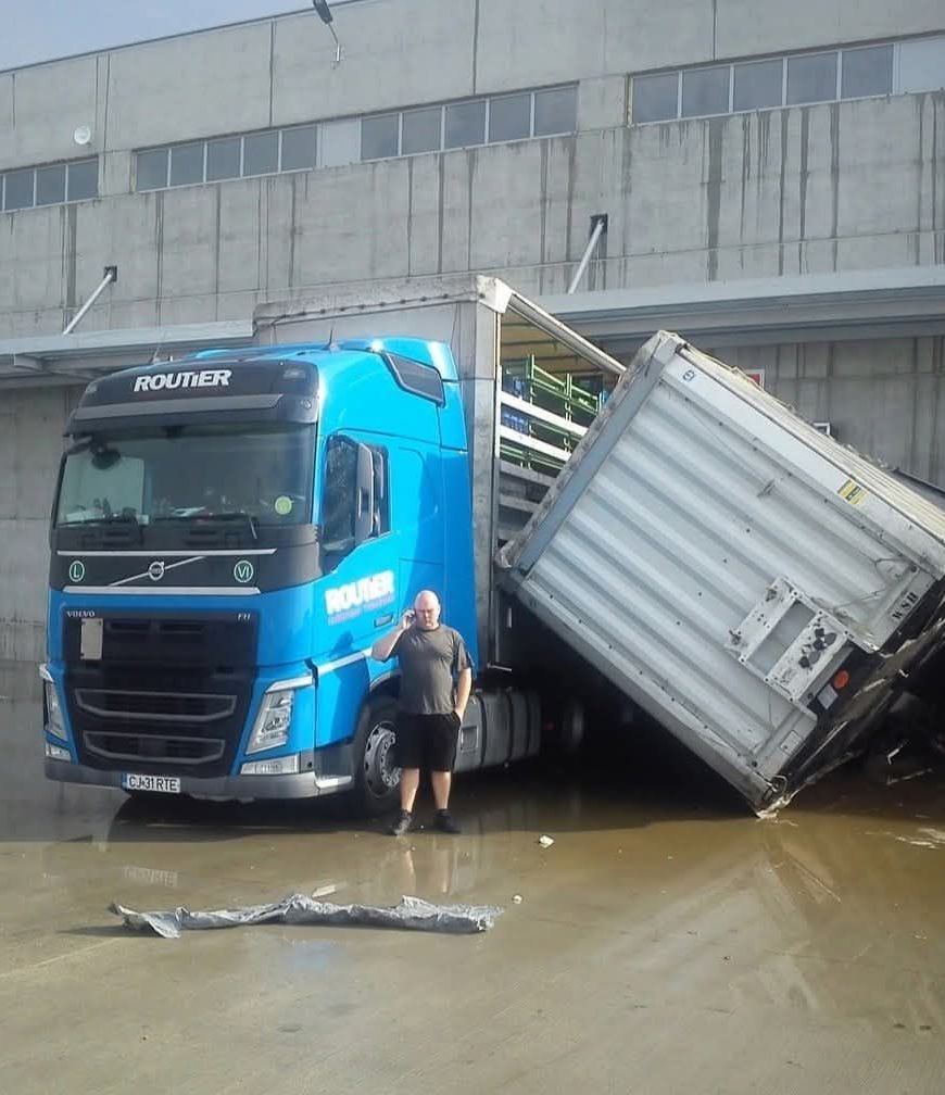 Kamión s modrou kabínou pri nehode pri sklade, vodič stojí vedľa nakloneného prívesu na mokrej zemi.
