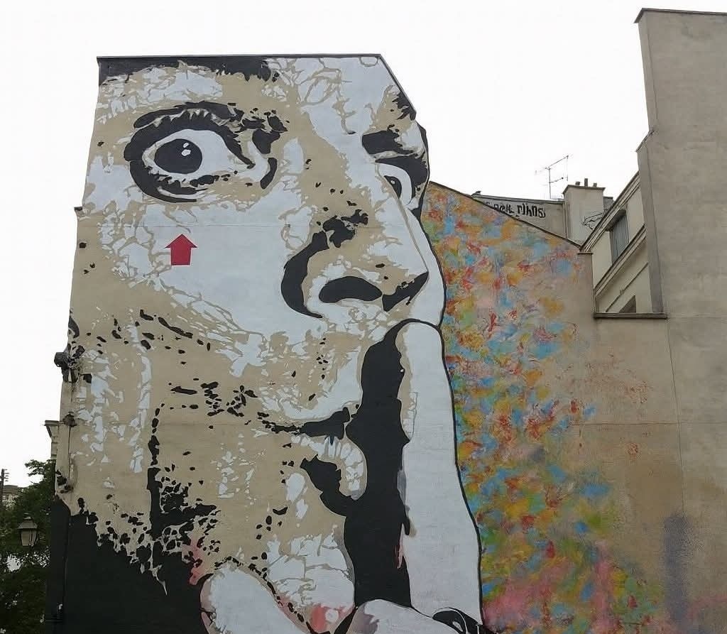 Nástenná maľba veľkej tváre s prstom na perách, symbolizujúca ticho na farebnom pozadí.