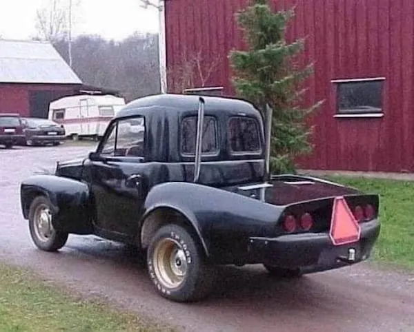 Unikátne čierne auto s upravenou karosériou, trojuholníkovým výstražným znakom vzadu, zaparkované pred červeným domom.