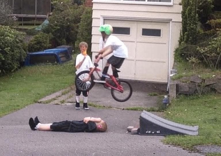 Chlapec na bicykli skáče cez ležiaceho kamaráta na príjazdovej ceste pred garážou.