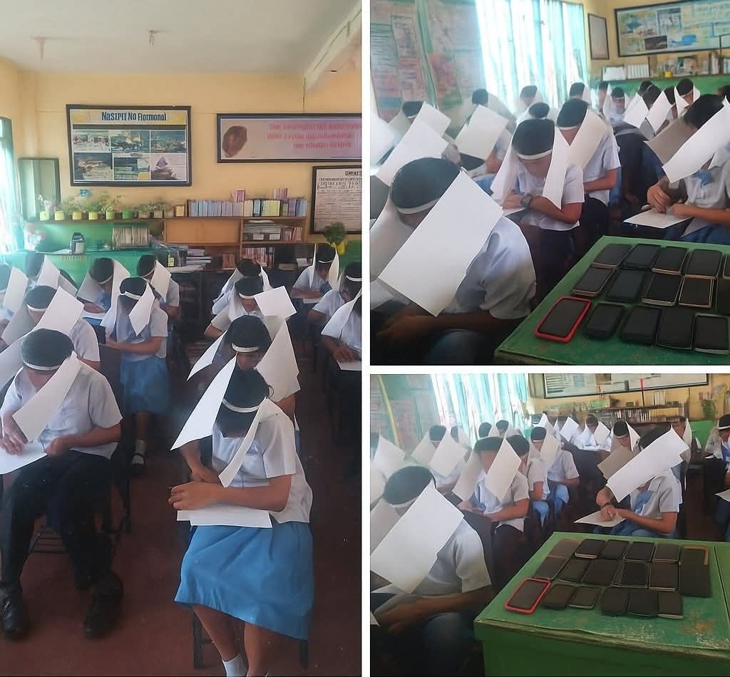 Študenti v triede nosia papiere okolo hlavy počas písomky, stoly sú plné telefónov.