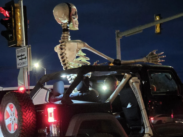 Obrovská kostra sediaca v otvorenom džípe na nočnej ulici, osvetlená dopravným svetlom na križovatke.