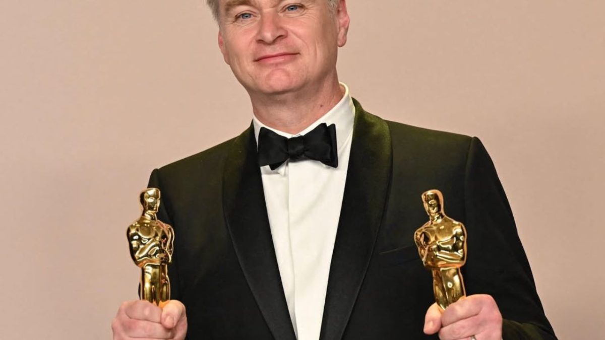 Osoba v smokingu drží dve zlaté sochy, ktoré vyzerajú ako ceny na slávnostnom ceremoniáli.