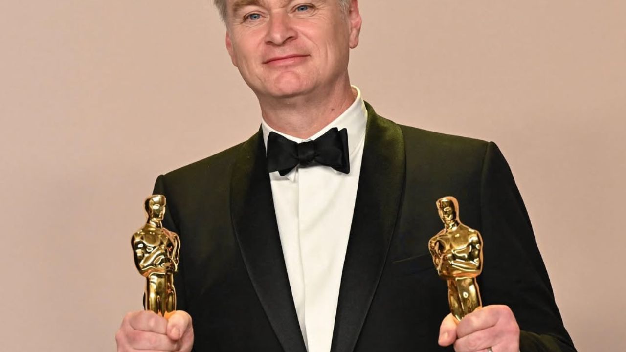 Osoba v smokingu drží dve zlaté sochy, ktoré vyzerajú ako ceny na slávnostnom ceremoniáli.